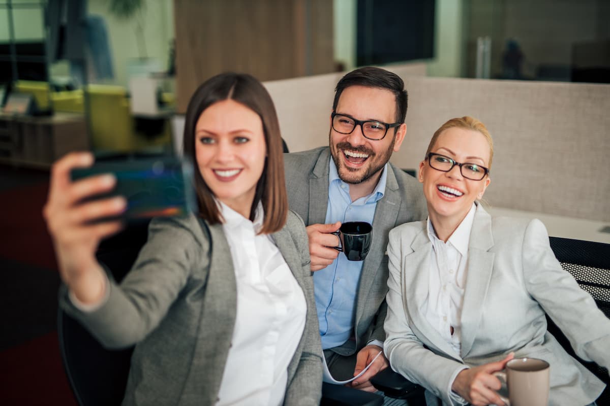 3 Arbeitskolleginnen und Kollegen machen auf der Arbeit ein Selfie und lächeln in die Kamera